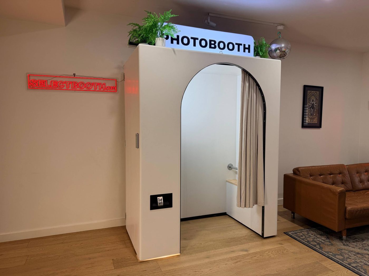 The Select Booth photo booth, a white arched booth with a lit PHOTOBOOTH sign on top, standing against a wall beside a red SELECTBOOTH.com neon.